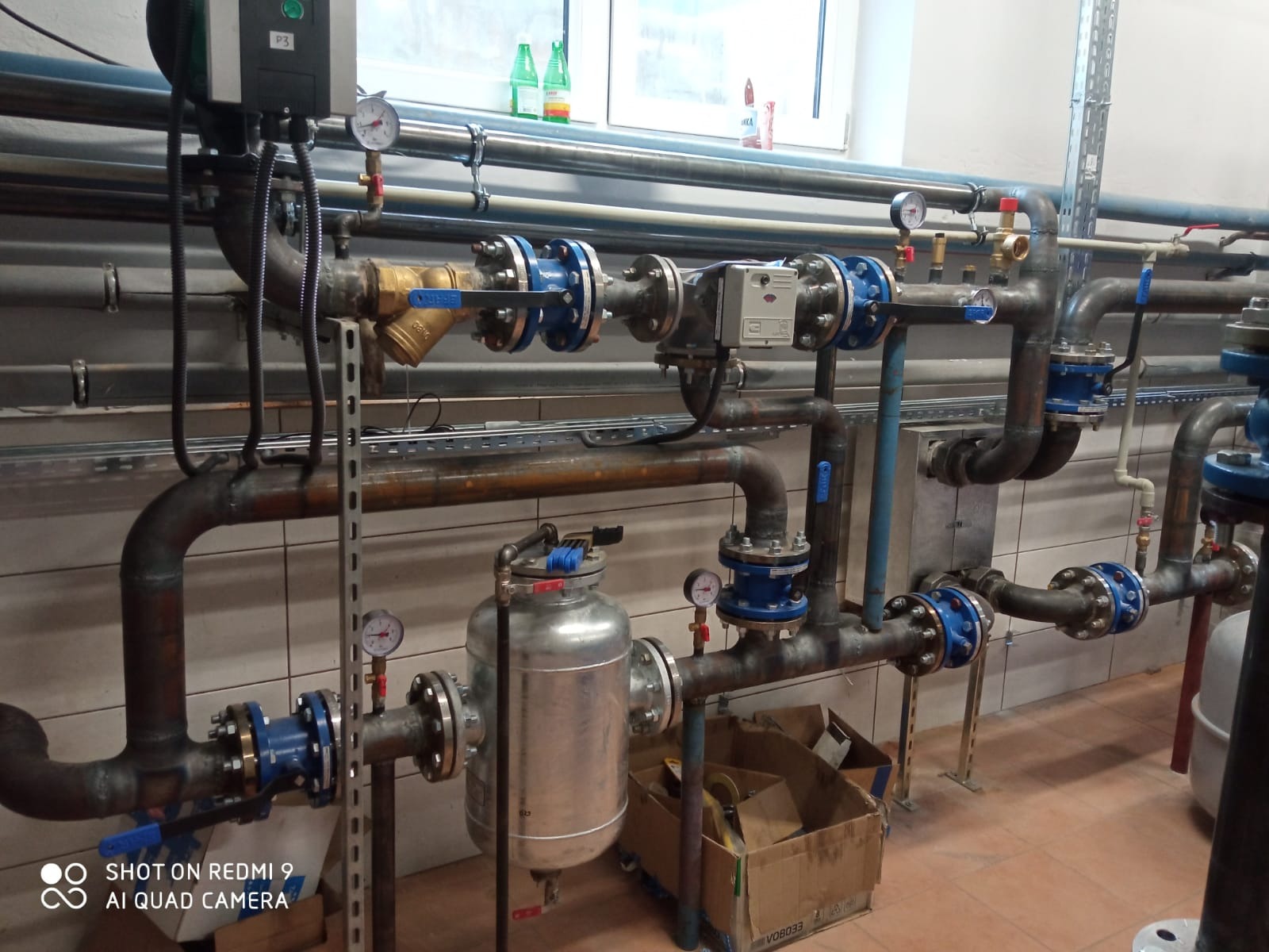 Złożony system rur z manometrami, zaworami z niebieskimi kołnierzami i filtrem, zainstalowany przy kafelkowanej ścianie, widoczne butelki z wodą na parapecie okna w tle.