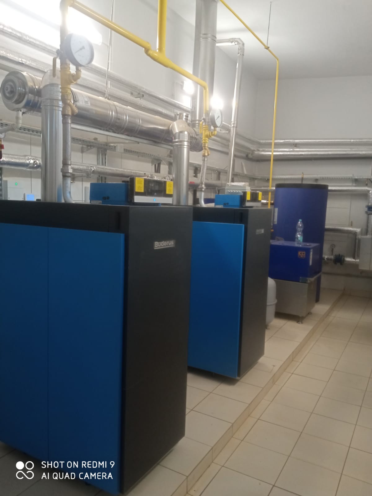 Kotłownia w Janowie Lubelskim: rzędy niebiesko-czarnych kotłów grzewczych Buderus z widocznymi rurami instalacji. Na jednym z kotłów butelka wody.