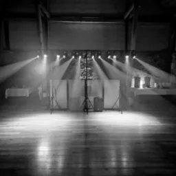 Czarno-białe zdjęcie pustej sceny z oświetleniem punktowym, drewniana podłoga, belki stropowe, przygotowana do występu.