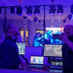 DJ uśmiechnięty i robiący zdjęcie telefonem, w tle parkiet z bawiącymi się ludźmi, oświetlenie dyskotekowe i balony, widoczne głośniki i monitory z oprogramowaniem DJ-skim