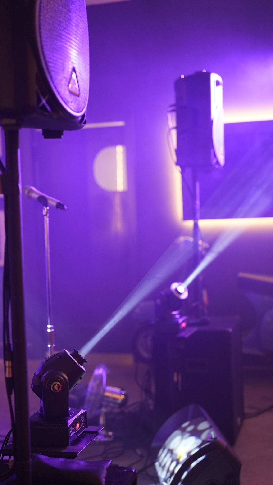 Scena oświetlona fioletowym światłem z widocznymi reflektorami, mikrofonem na statywie i głośnikami, tworząca atmosferę koncertową.
