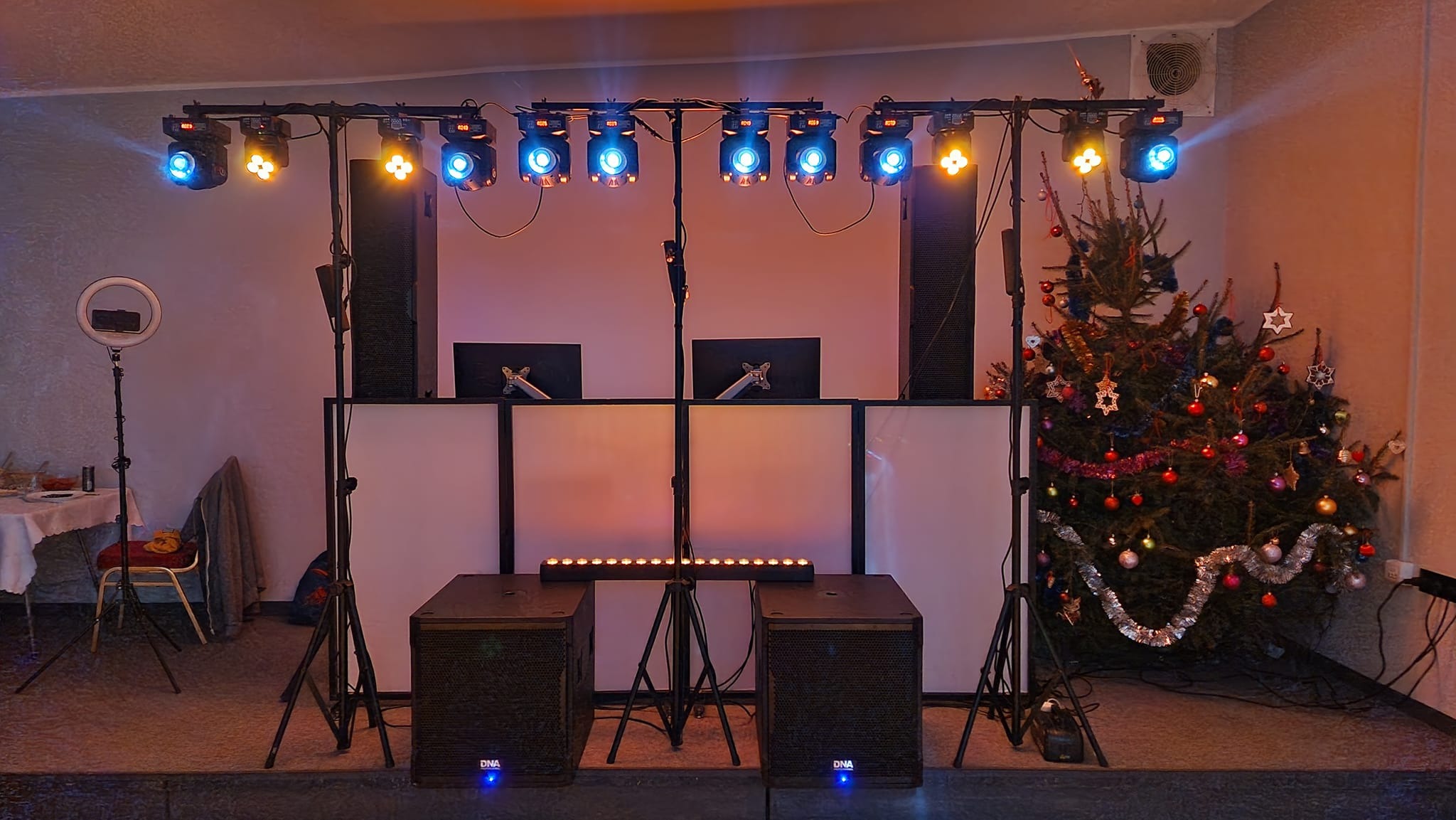 Scena DJ-a z oświetleniem LED w kolorach niebieskim i żółtym, z choinką świąteczną po prawej stronie, ring light po lewej stronie, dwa monitory na białym panelu i dwa subwoofery na podłodze.