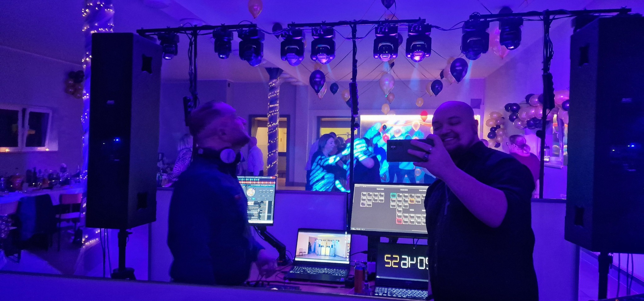 DJ uśmiechnięty i robiący zdjęcie telefonem, w tle parkiet z bawiącymi się ludźmi, oświetlenie dyskotekowe i balony, widoczne głośniki i monitory z oprogramowaniem DJ-skim