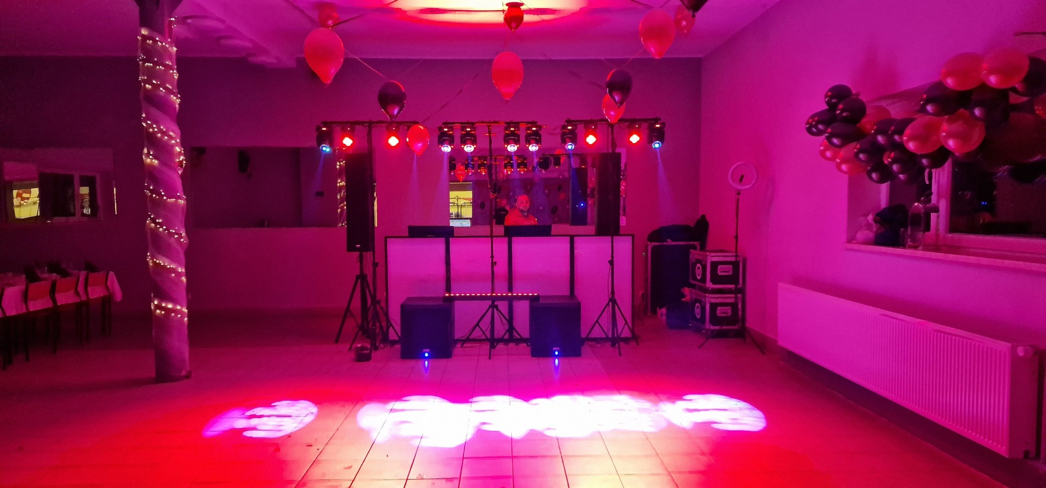 Sala udekorowana balonami z DJ-em za konsoletą, oświetlona czerwonym światłem, kolumna owinięta światełkami, widoczne głośniki i reflektory.