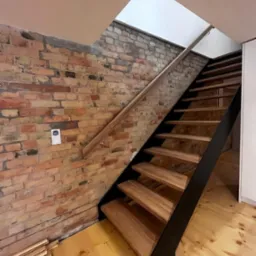 Nowoczesne schody z drewnianymi stopniami i czarną metalową konstrukcją boczną, zamontowane przy ścianie z czerwonej cegły, z drewnianą poręczą i widocznym fragmentem drewnianej podłogi.