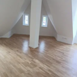 Poddasze po remoncie z jasną drewnianą podłogą ułożoną w jodełkę, białe ściany i słup konstrukcyjny pośrodku, dwa okna dachowe.