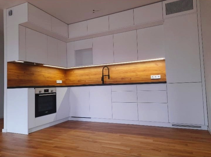Nowoczesna, jasna kuchnia z białymi szafkami na wymiar, drewnianym blatem i podświetleniem LED, widziana z niskiego kąta, ukazująca parkiet na podłodze.