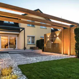 Nowoczesna drewniana konstrukcja z oświetleniem punktowym w ogrodzie, z elementami dekoracyjnymi i ścieżką z kamieni.