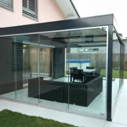 Zadaszony taras z przesuwnymi szklanymi ścianami, wyposażony w meble ogrodowe z ciemnego rattanu i stół z krzesłami, widok na trawnik i sąsiedni dom.