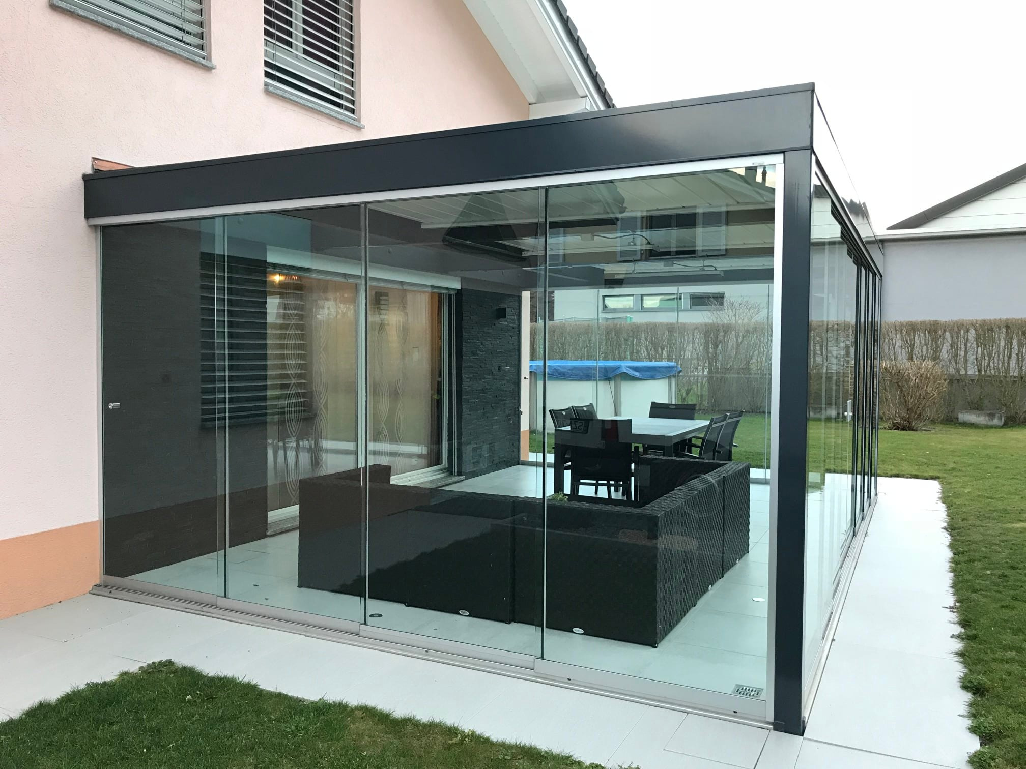 Zadaszony taras z przesuwnymi szklanymi ścianami, wyposażony w meble ogrodowe z ciemnego rattanu i stół z krzesłami, widok na trawnik i sąsiedni dom.