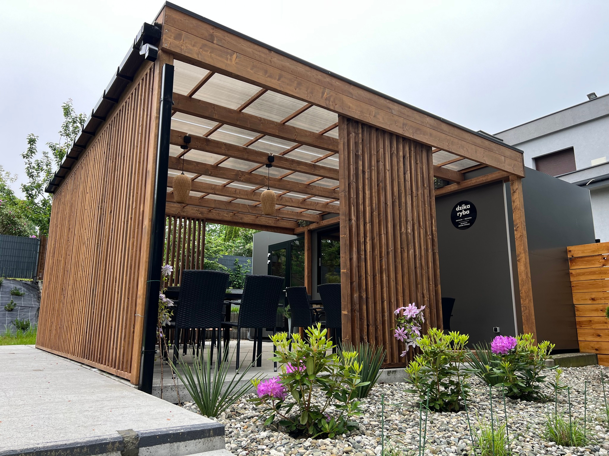 Nowoczesna drewniana konstrukcja z przesuwanymi panelami, prawdopodobnie altana lub pergola, z przezroczystym zadaszeniem, oświetleniem i czarnymi meblami ogrodowymi w środku, otoczona kamieniami...