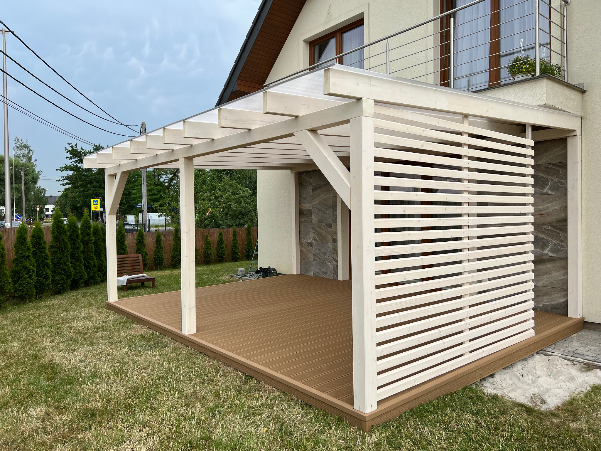 Biała konstrukcja z drewnianymi lamelami i przezroczystym dachem, przylegająca do domu z balkonem, z brązową podłogą na trawniku.