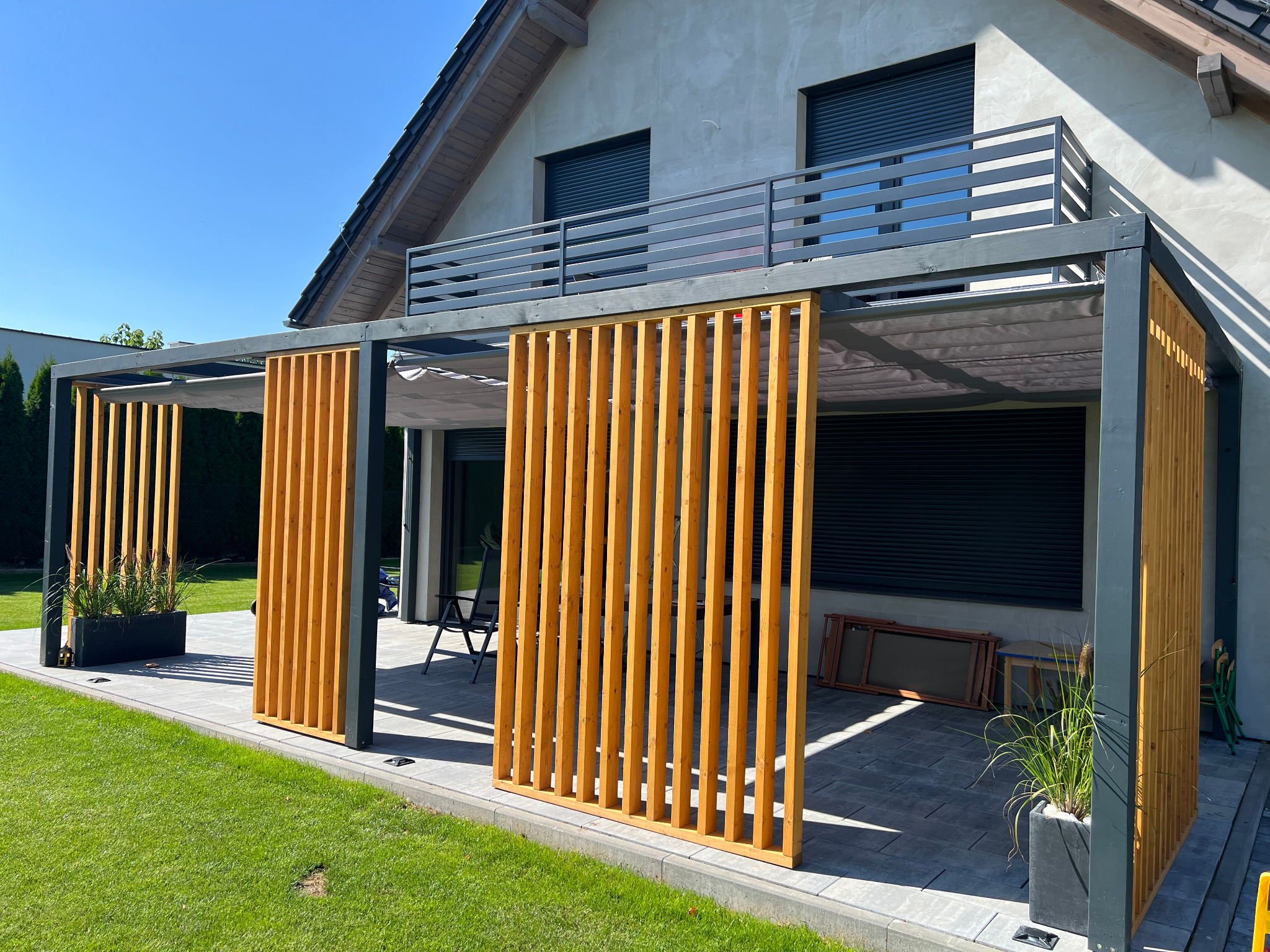 Nowoczesna pergola z drewnianymi panelami lamelowymi, szarą konstrukcją i rozsuwanym zadaszeniem, na tle domu z balkonem i zadbanym trawnikiem.