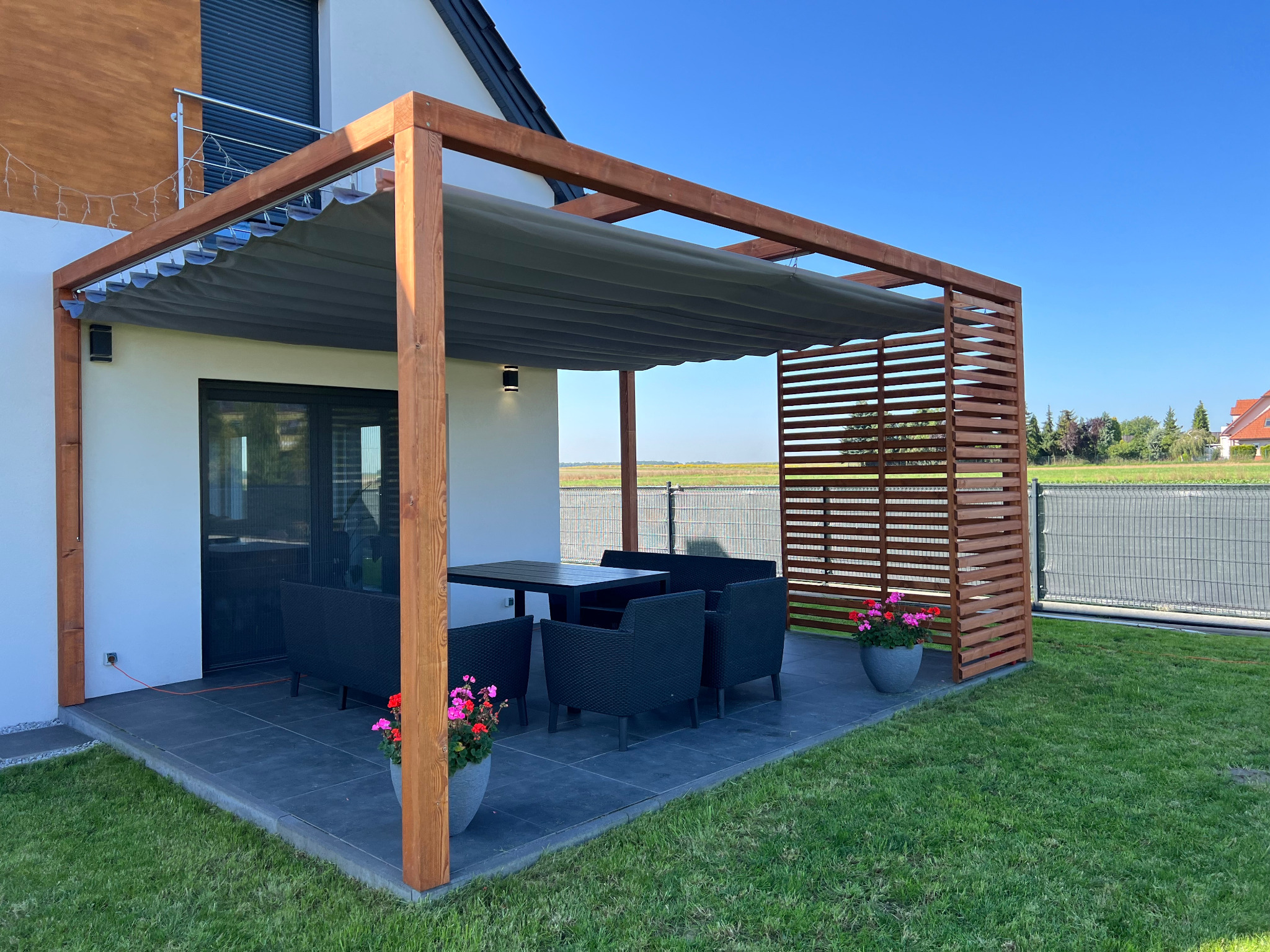 Nowoczesna pergola z drewnianą konstrukcją i rozsuwanym szarym zadaszeniem, wyposażona w czarne meble ogrodowe i donice z kwiatami, na tle zielonego trawnika i błękitnego nieba.