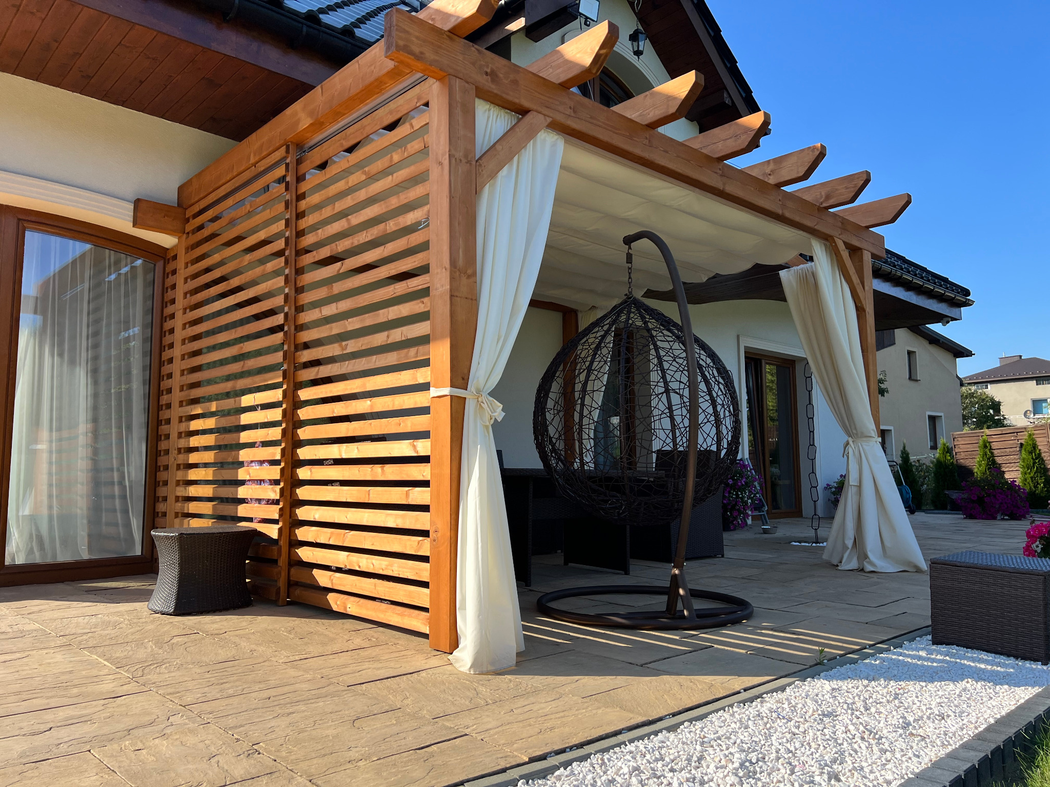 Drewniana pergola z lamelami i białymi zasłonami, wiszące krzesło, taras z płyt kamiennych i białego żwiru w słoneczny dzień.