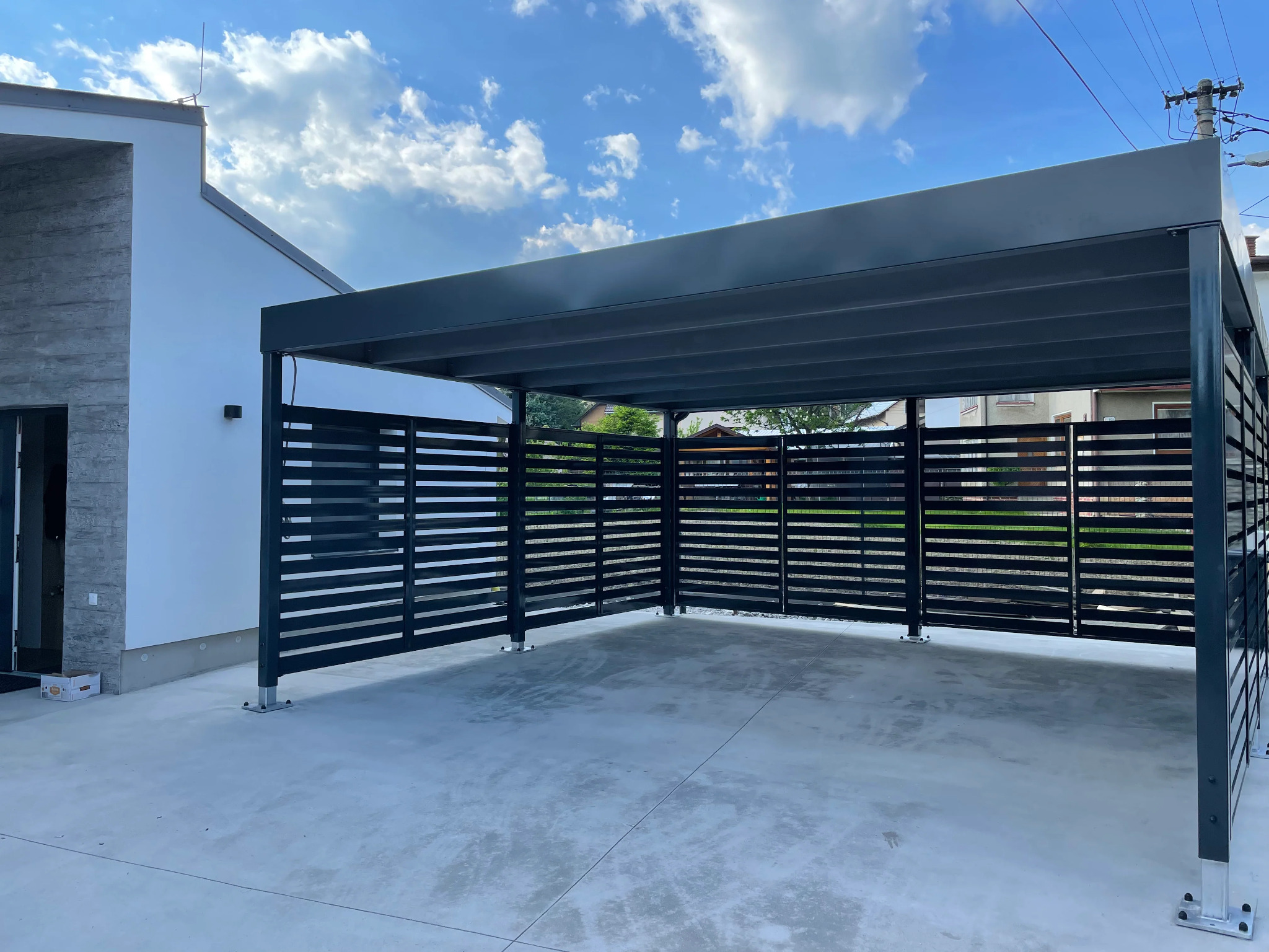 Nowoczesna, czarna pergola z poziomymi deskami, osłaniająca betonowy podjazd przed domem z białymi ścianami i błękitnym niebem w tle.