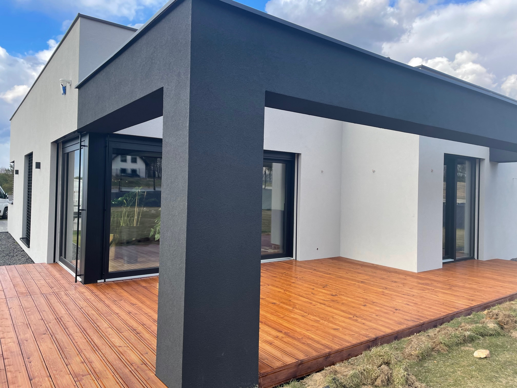 Nowoczesny taras z drewnianą deską, przylegający do białego budynku z czarnymi elementami konstrukcyjnymi i dużymi oknami, widok z zewnątrz.