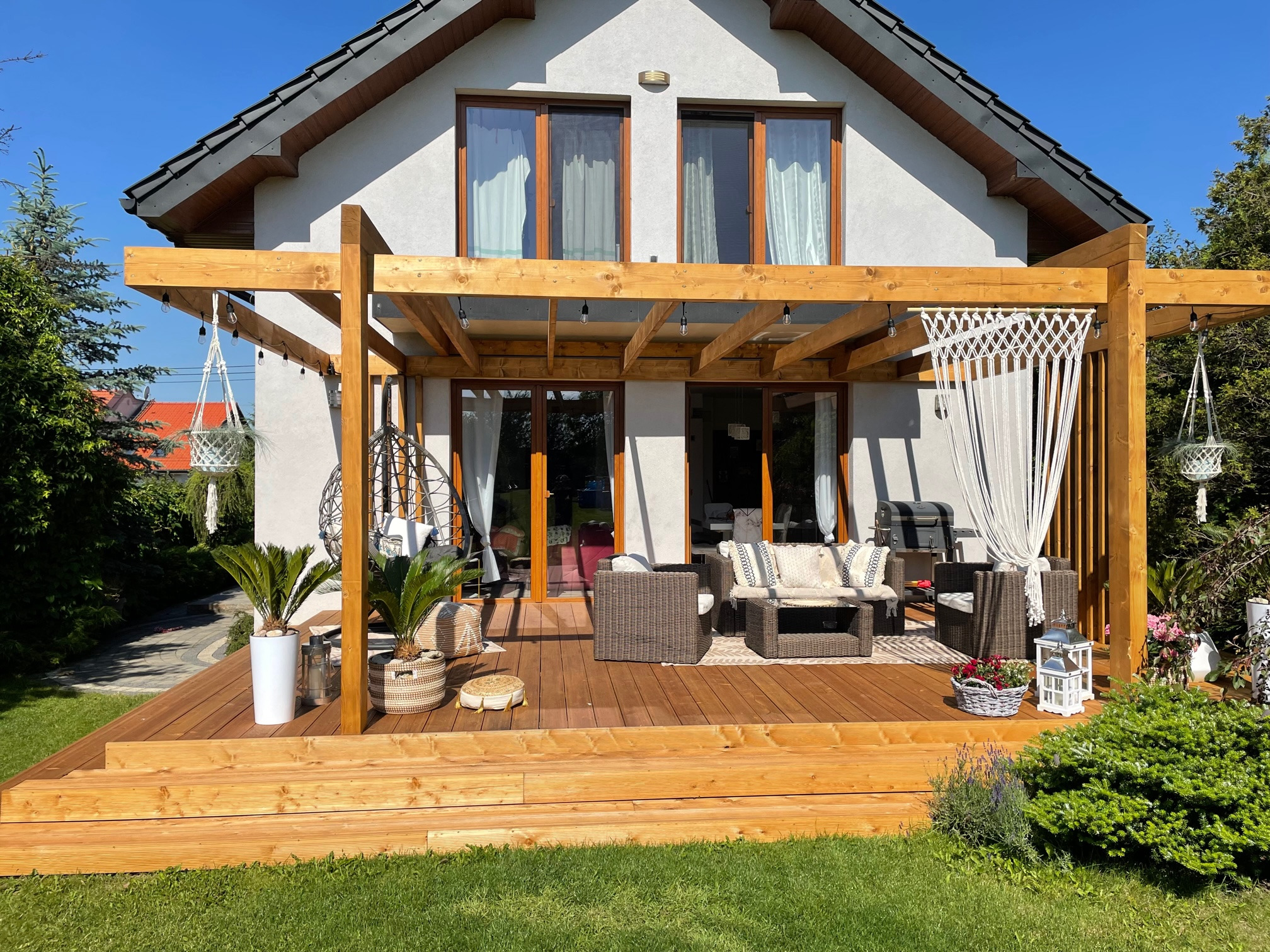 Drewniany taras z pergolą przy domu, wyposażony w meble ogrodowe, wiszące dekoracje i rośliny w donicach, oświetlony girlandami żarówek.