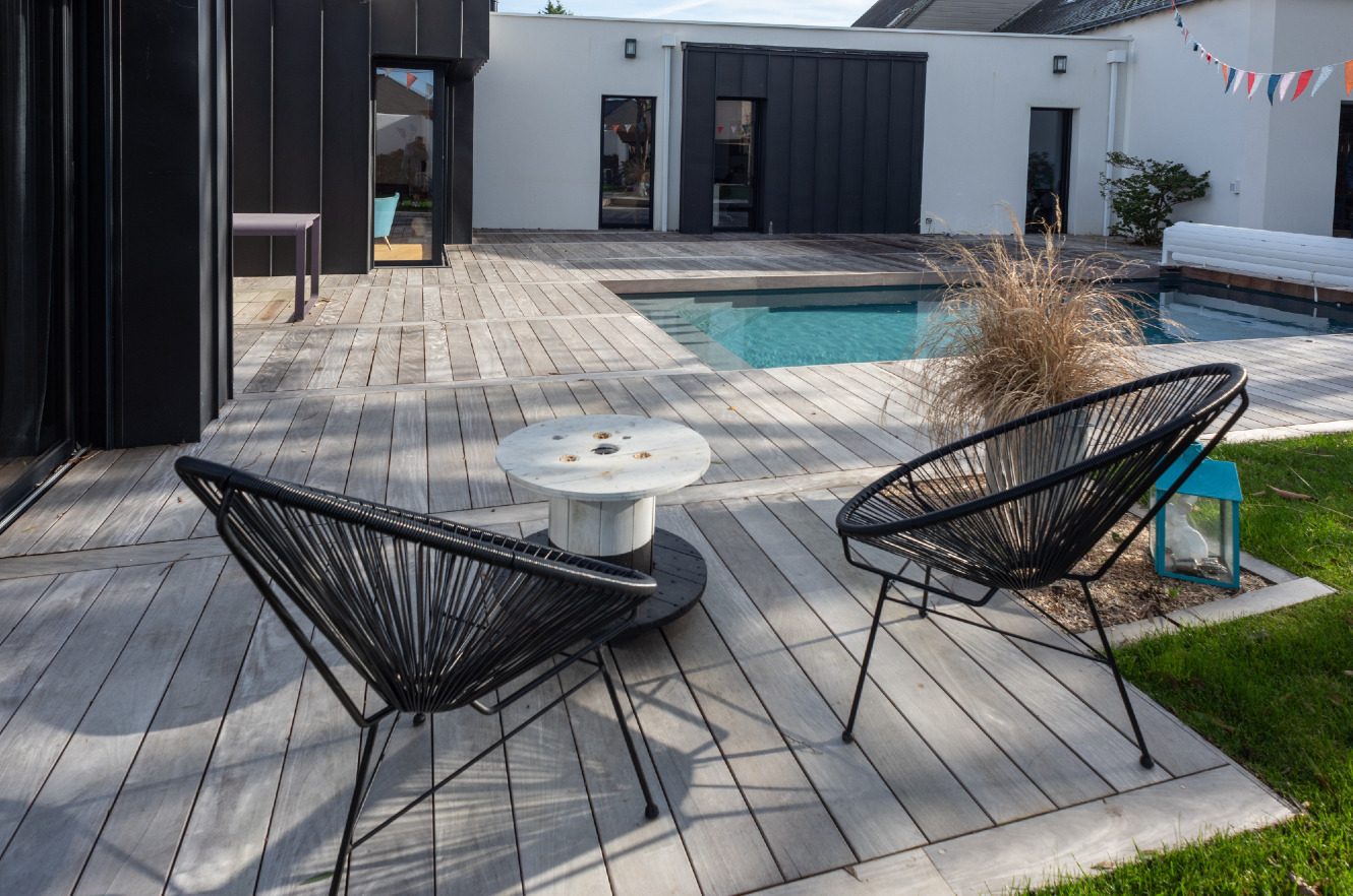 Jasnoszary taras z desek z wbudowanym basenem, dwoma czarnymi krzesłami sznurkowymi i stolikiem z drewnianej szpuli po kablach, nowoczesny dom z czarno-białą elewacją w tle.