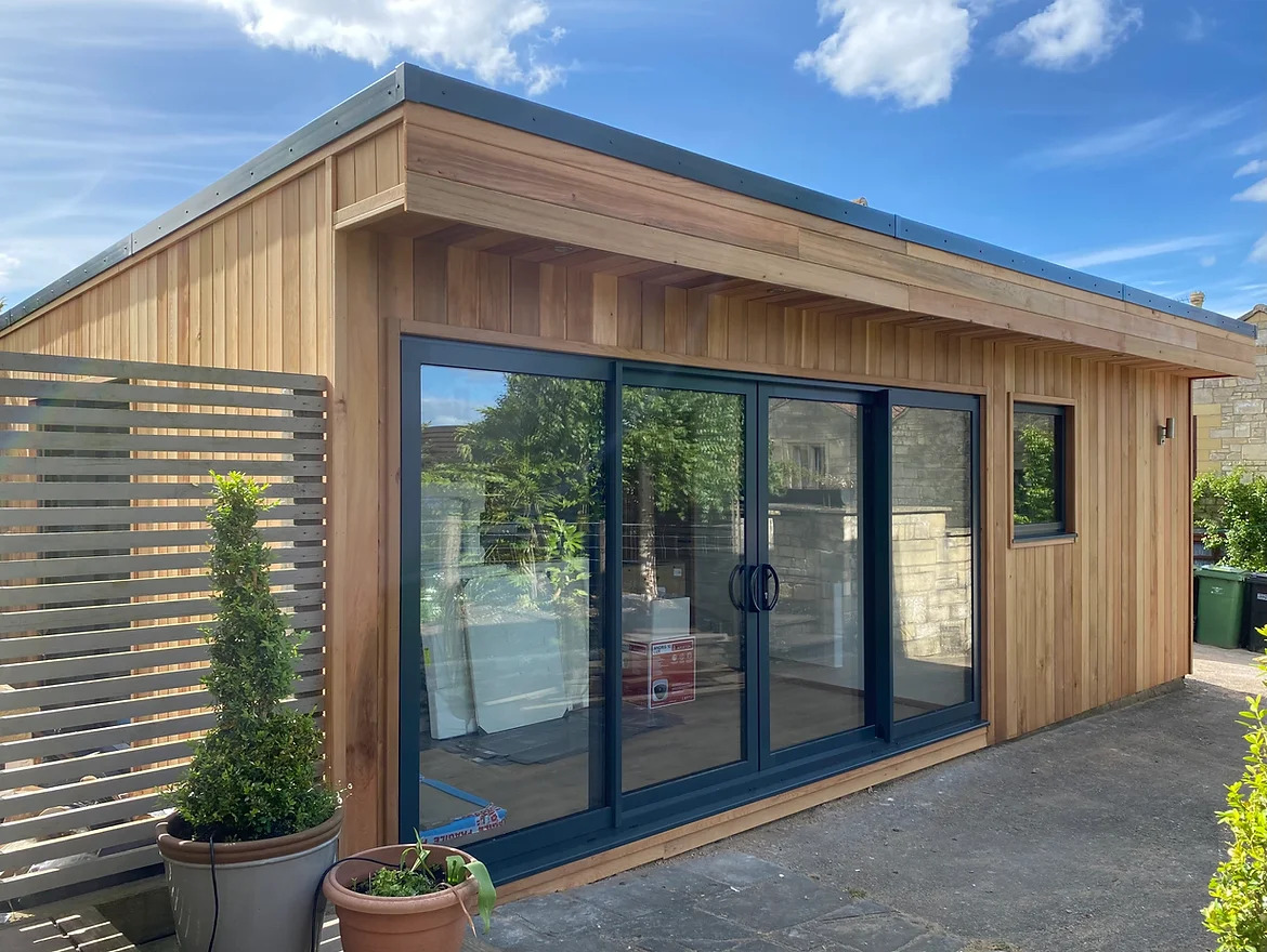 Nowoczesna, drewniana konstrukcja z dużymi przesuwnymi drzwiami szklanymi, wykończona drewnianą boazerią i ciemnoszarą listwą dachową, otoczona brukowanym patio i roślinnością.