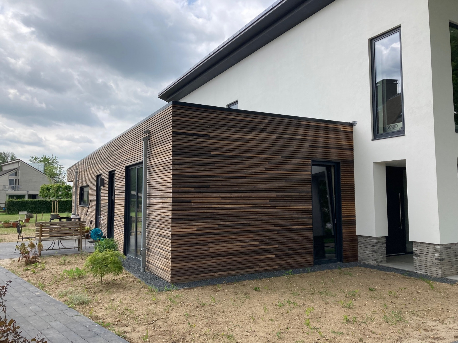 Nowoczesny dom z elewacją z poziomych desek w odcieniach brązu, kontrastujący z białą częścią budynku i czarnymi ramami okiennymi, otoczony minimalistycznym ogrodem z młodymi roślinami.