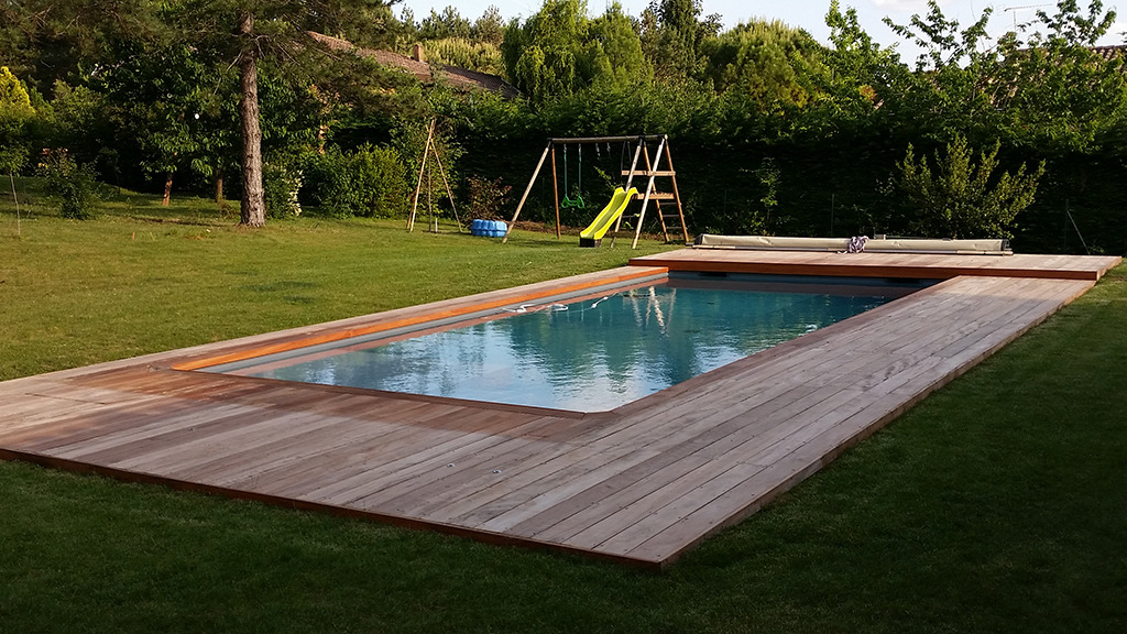 Drewniany taras otaczający basen w ogrodzie, z placem zabaw w tle, widok z poziomu ziemi.