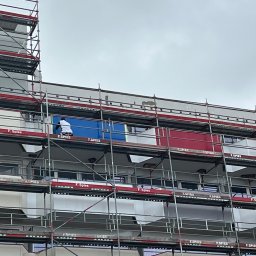 WOJBUD Plus Wojciech Budnik - Pracownik w białej koszulce maluje fragment elewacji budynku z rusztowania na kolor niebieski, obok pomalowany fragment na kolor czerwony, pochmurne niebo w tle.