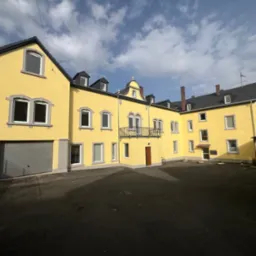 Żółty budynek z szarymi oknami i dachem, widok z dziedzińca w słoneczny dzień, z widocznymi detalami architektonicznymi i garażem po lewej stronie.