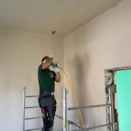 Mężczyzna na rusztowaniu, w masce i rękawicach, maluje sufit w pomieszczeniu za pomocą pistoletu natryskowego z żółtym przewodem, widoczne narzędzia w pasie.