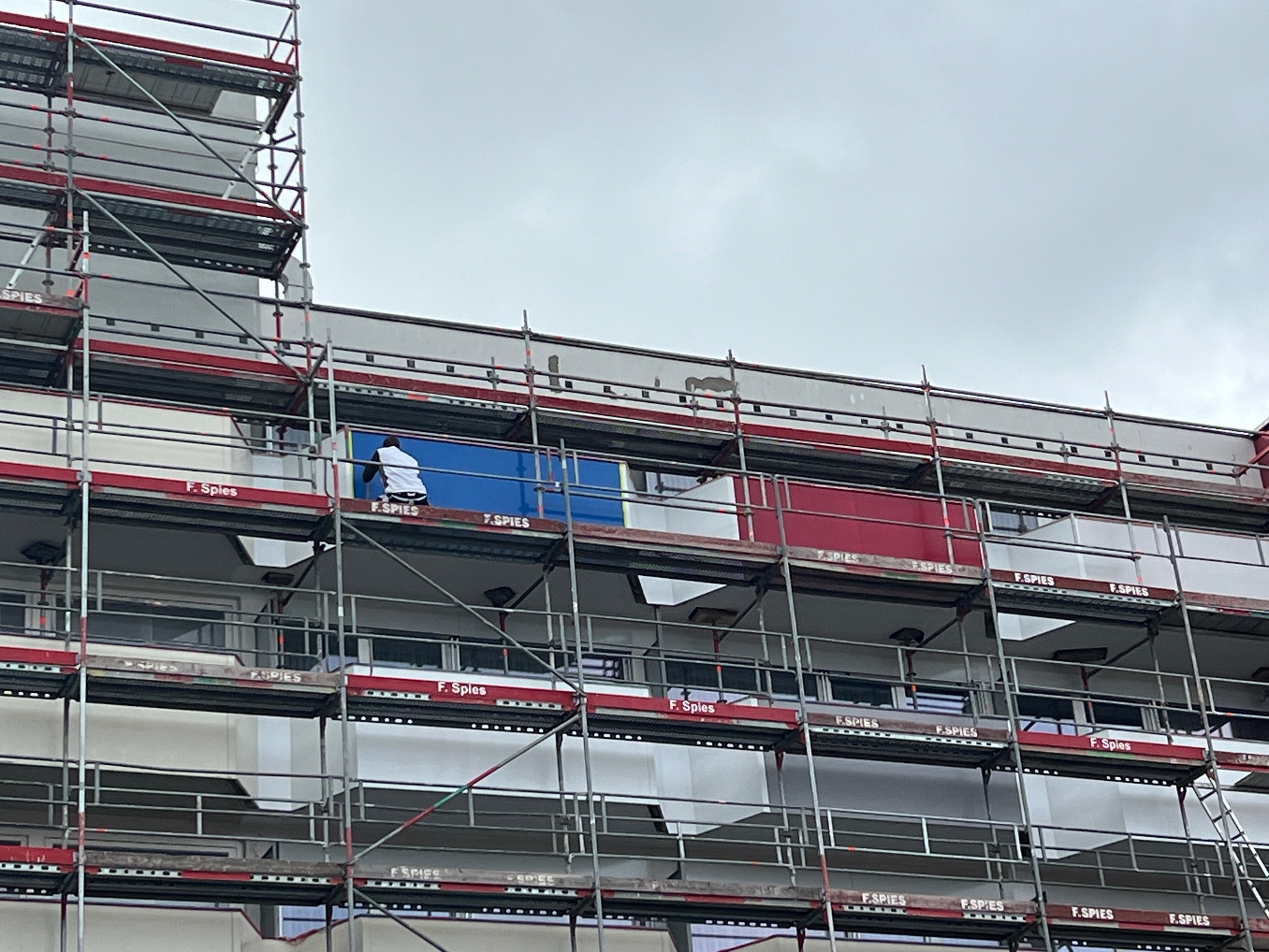 Pracownik w białej koszulce maluje fragment elewacji budynku z rusztowania na kolor niebieski, obok pomalowany fragment na kolor czerwony, pochmurne niebo w tle.