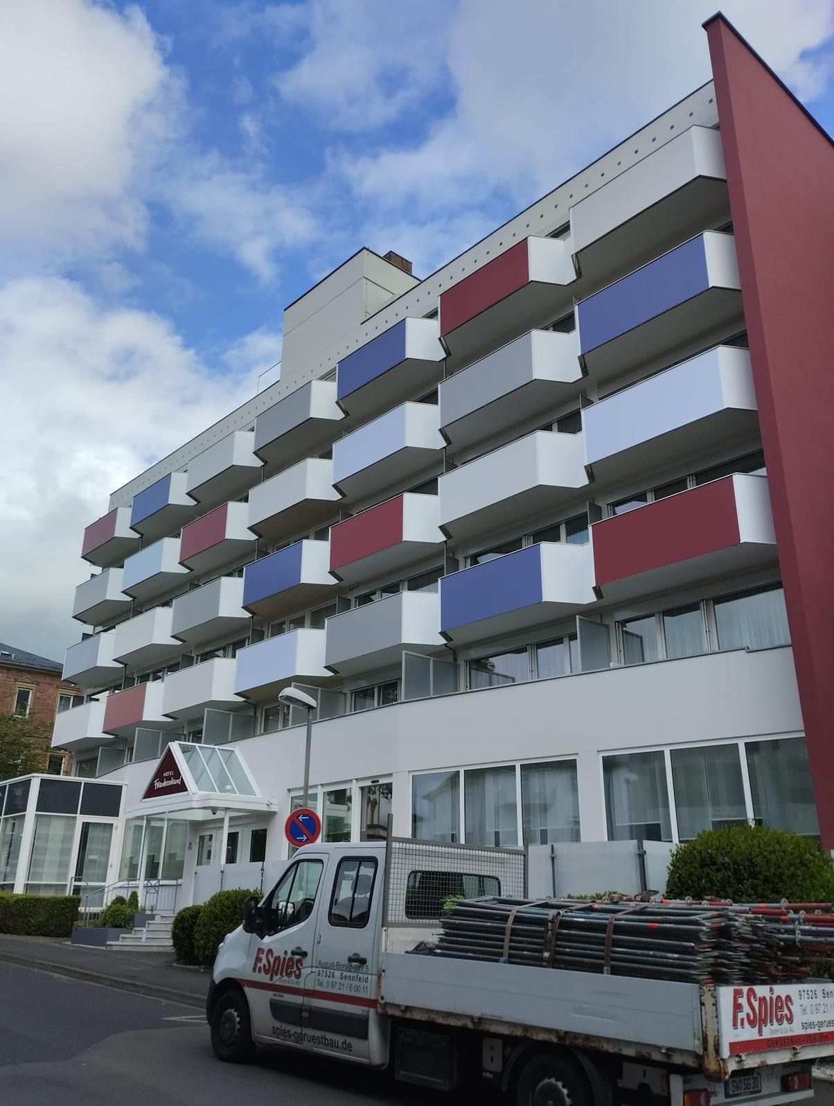 Wielopiętrowy budynek hotelowy z balkonami pomalowanymi na biało, czerwono i niebiesko. Na pierwszym planie samochód dostawczy z rusztowaniem.