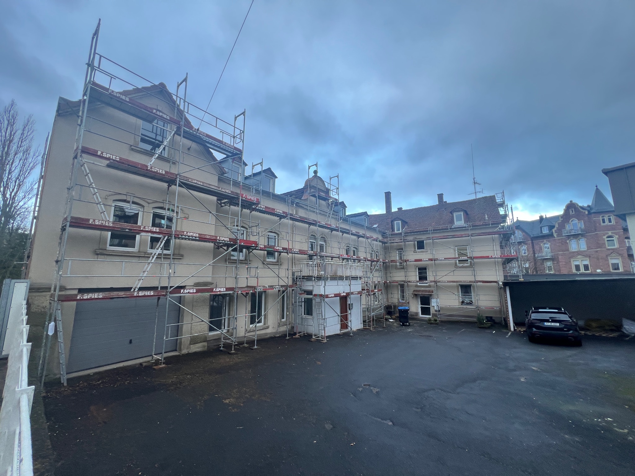 Budynek wielorodzinny w trakcie remontu elewacji, otoczony rusztowaniami z widocznym logo firmy F. SPIES, na pierwszym planie mokry asfalt parkingu i zaparkowany czarny samochód.