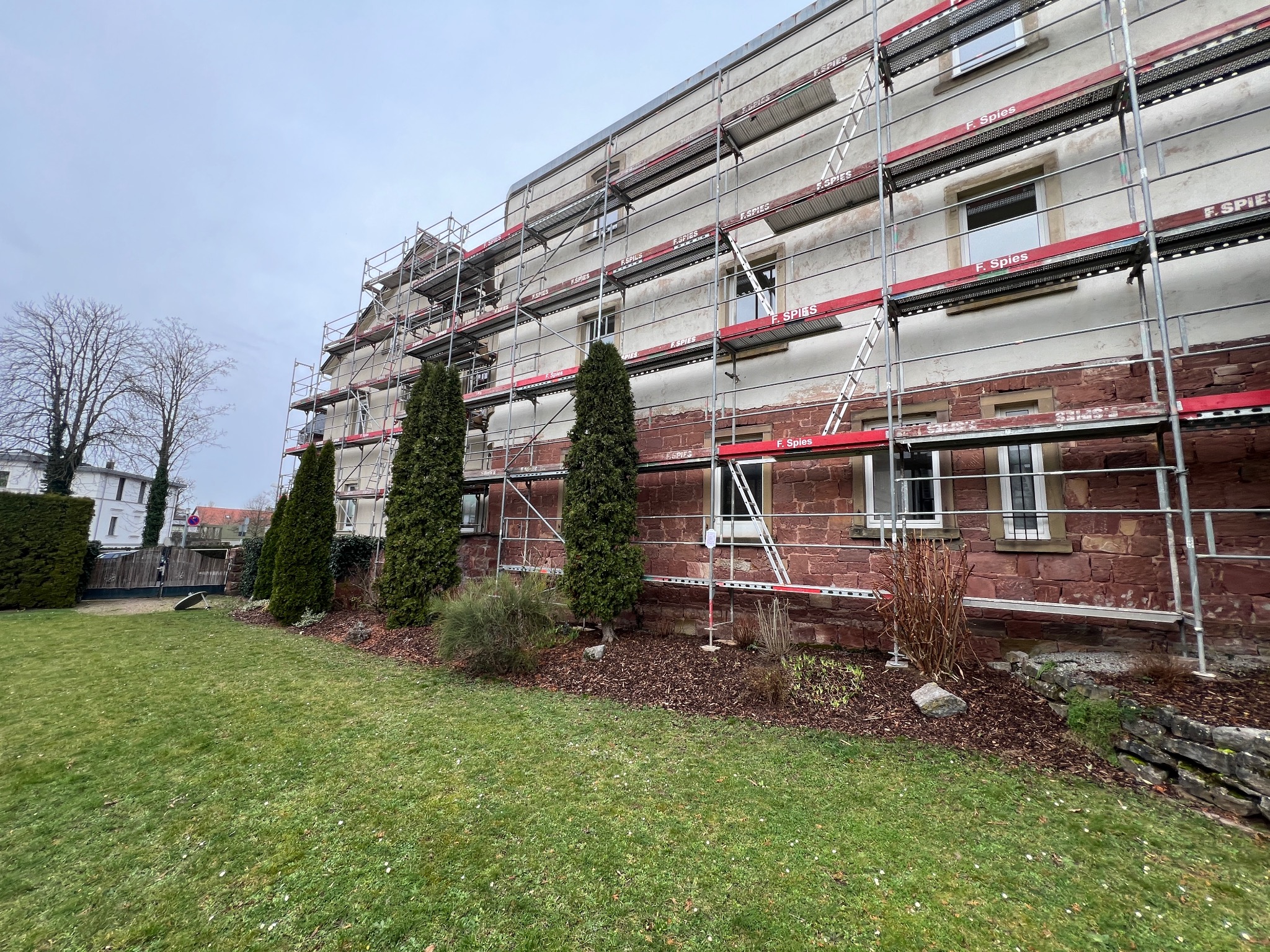 Wielopiętrowy budynek w trakcie prac elewacyjnych, otoczony rusztowaniami z widocznymi oznaczeniami firmy, z zadbanym trawnikiem i zielenią w tle, w pochmurny dzień.