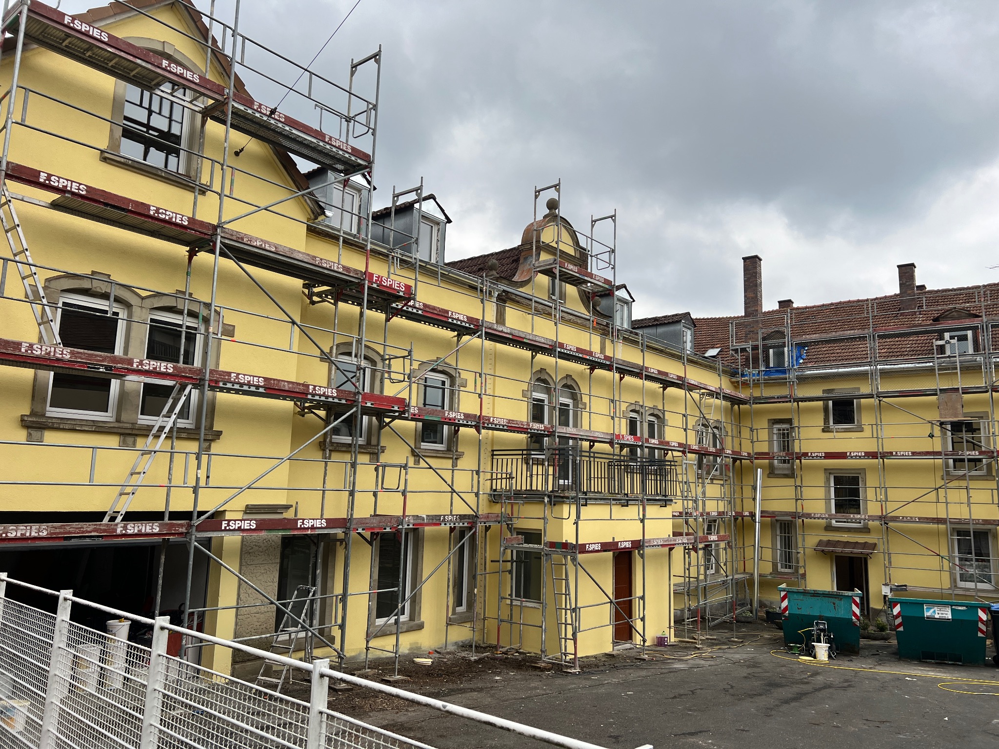 Żółta elewacja budynku w trakcie prac remontowych, otoczona rusztowaniami z widocznym logo 'F.SPIES', dwa kontenery na śmieci i sprzęt malarski na dziedzińcu, pochmurne niebo w tle.