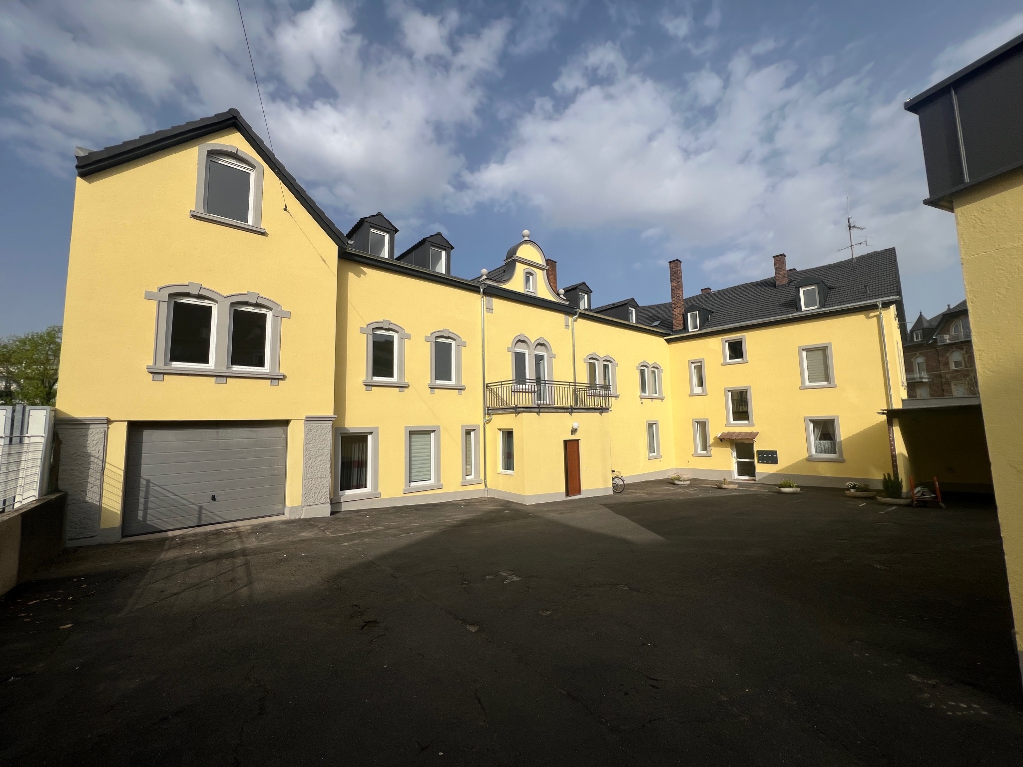 Żółty budynek z szarymi oknami i dachem, widok z dziedzińca w słoneczny dzień, z widocznymi detalami architektonicznymi i garażem po lewej stronie.