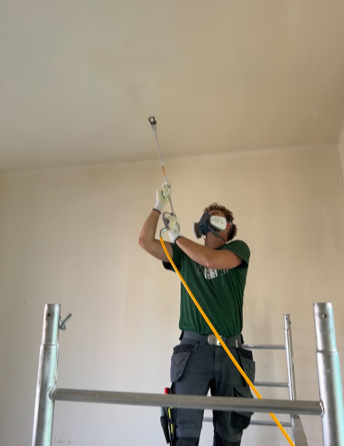 Malarz w masce ochronnej na rusztowaniu, maluje sufit beżową farbą za pomocą pistoletu natryskowego z długą lancą i żółtym wężem doprowadzającym farbę.