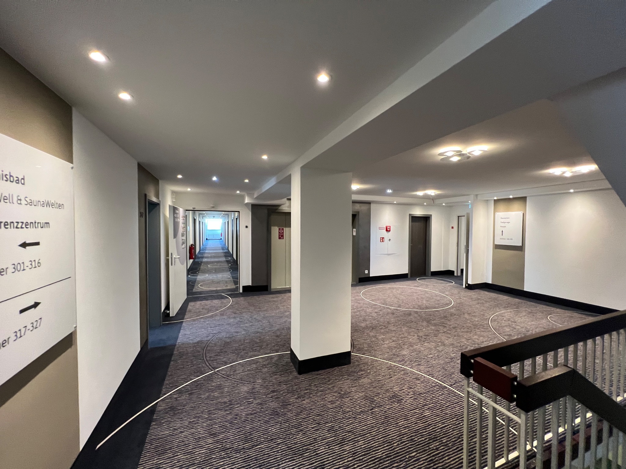 Długi korytarz hotelowy z wzorzystą wykładziną, białymi ścianami i sufitami z wbudowanymi lampami, widoczne oznaczenia kierunkowe oraz filar w centrum kadru.