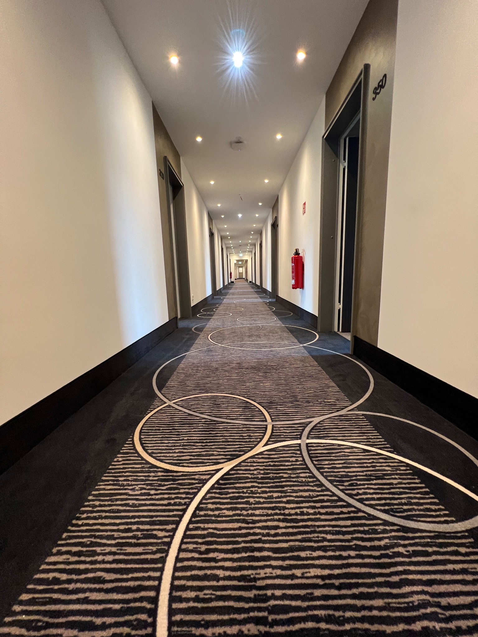 Długi korytarz hotelowy z wykładziną w paski i okręgi, numeracja pokoi nad drzwiami, białe ściany z szarą lamperią, widoczny hydrant po prawej stronie.