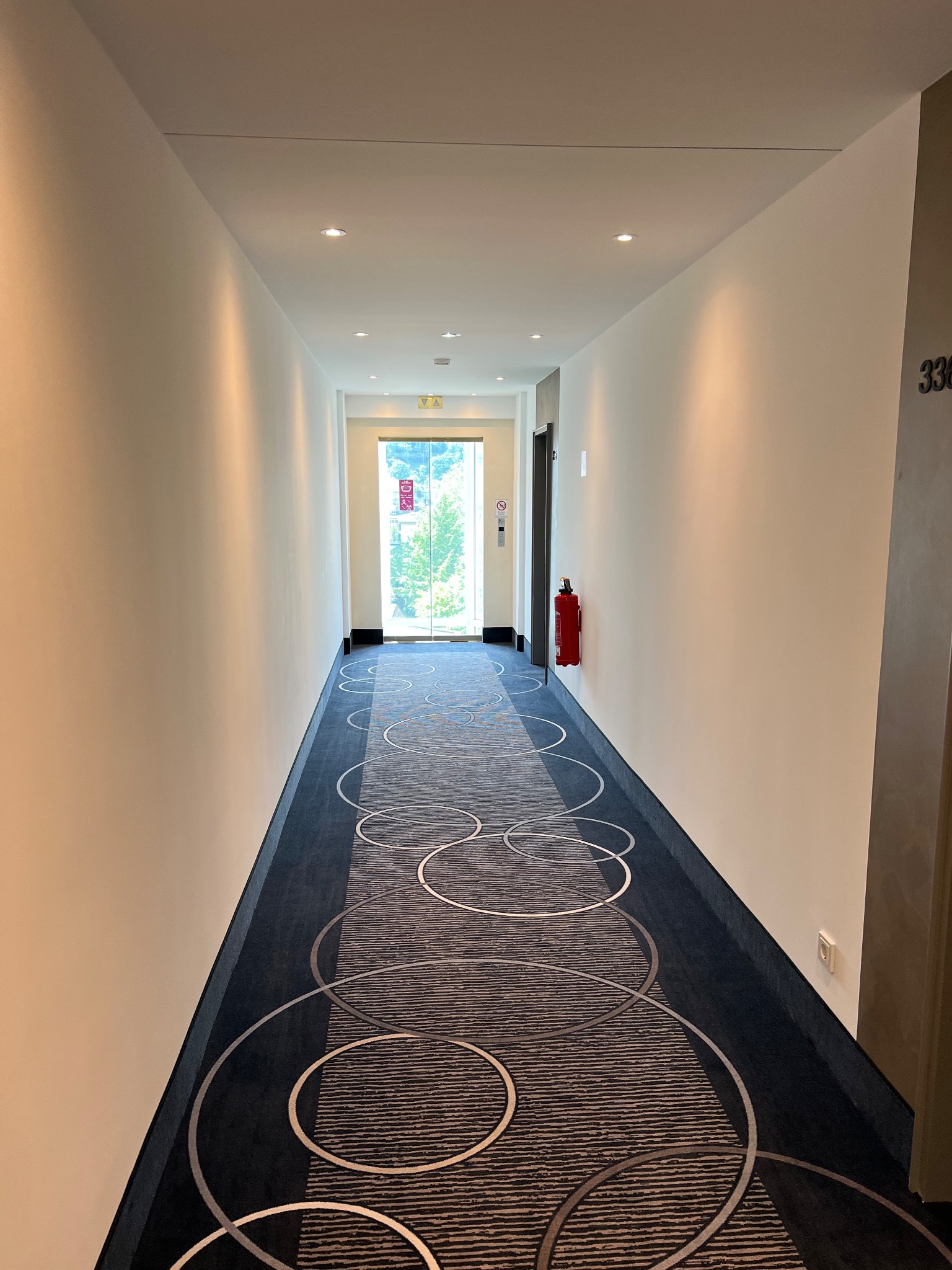 Długi korytarz hotelowy z kremowymi ścianami, wzorzystą wykładziną w odcieniach szarości i niebieskiego z motywem kół, zakończony przeszklonymi drzwiami z widokiem na zieleń. Na ścianie widoczna...