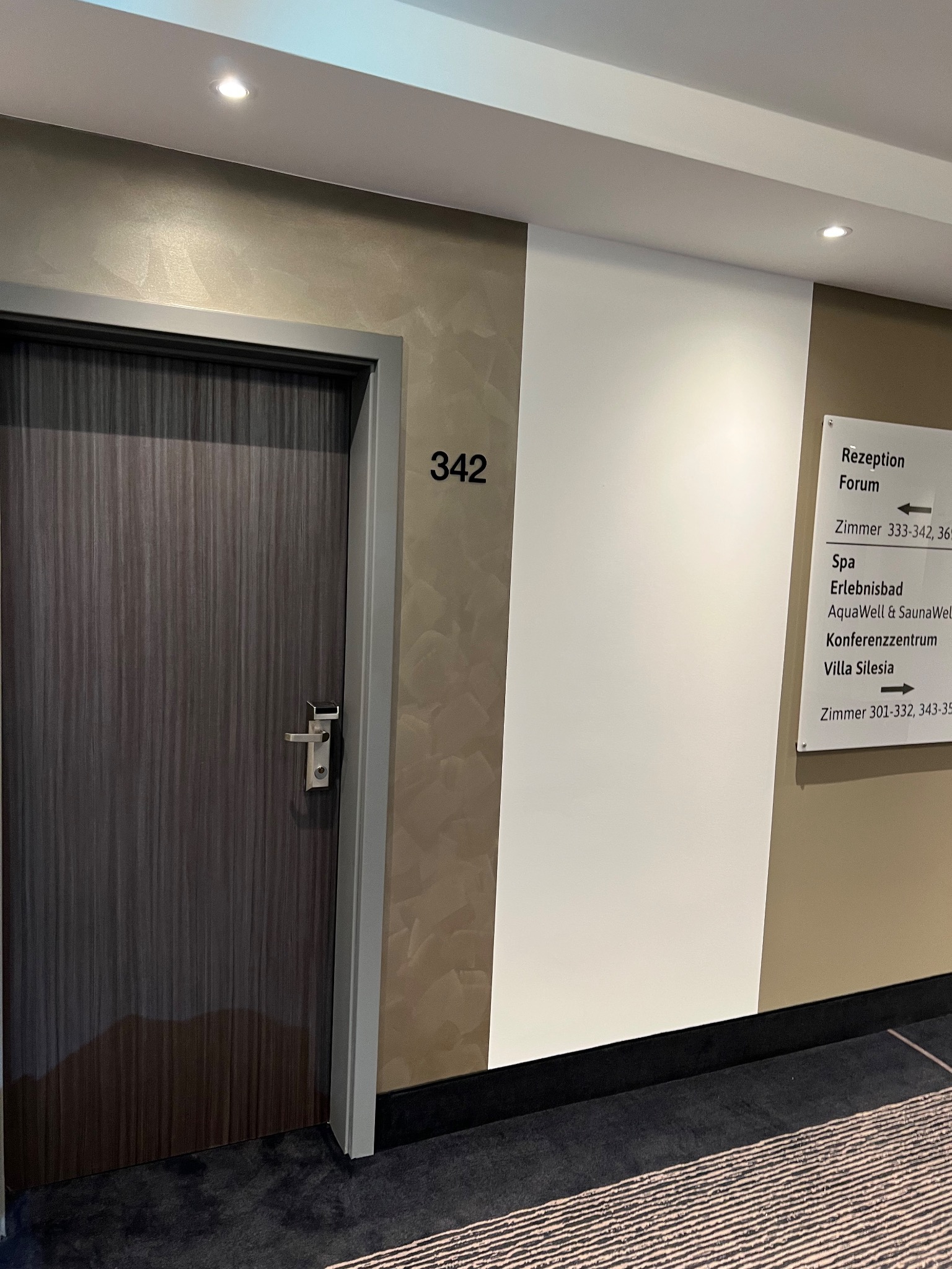 Korytarz hotelowy z ciemnymi drzwiami o numerze 342, ściana z dekoracyjnym malowaniem w odcieniach beżu i tabliczka informacyjna z napisem 'Rezeption Forum'.