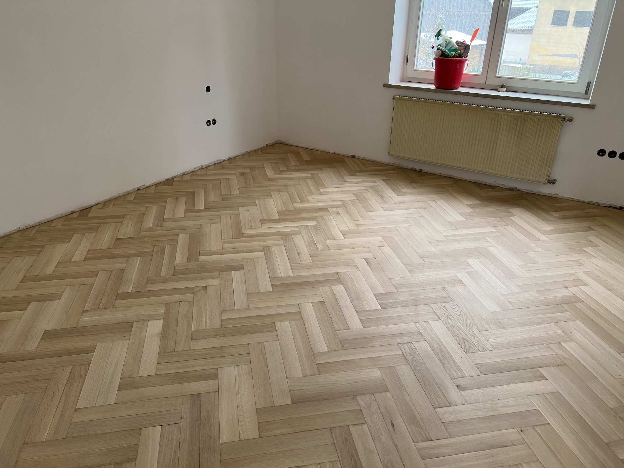 Jasne panele ułożone w jodełkę francuską w pustym pokoju z grzejnikiem pod oknem i puszkami elektrycznymi na ścianie.