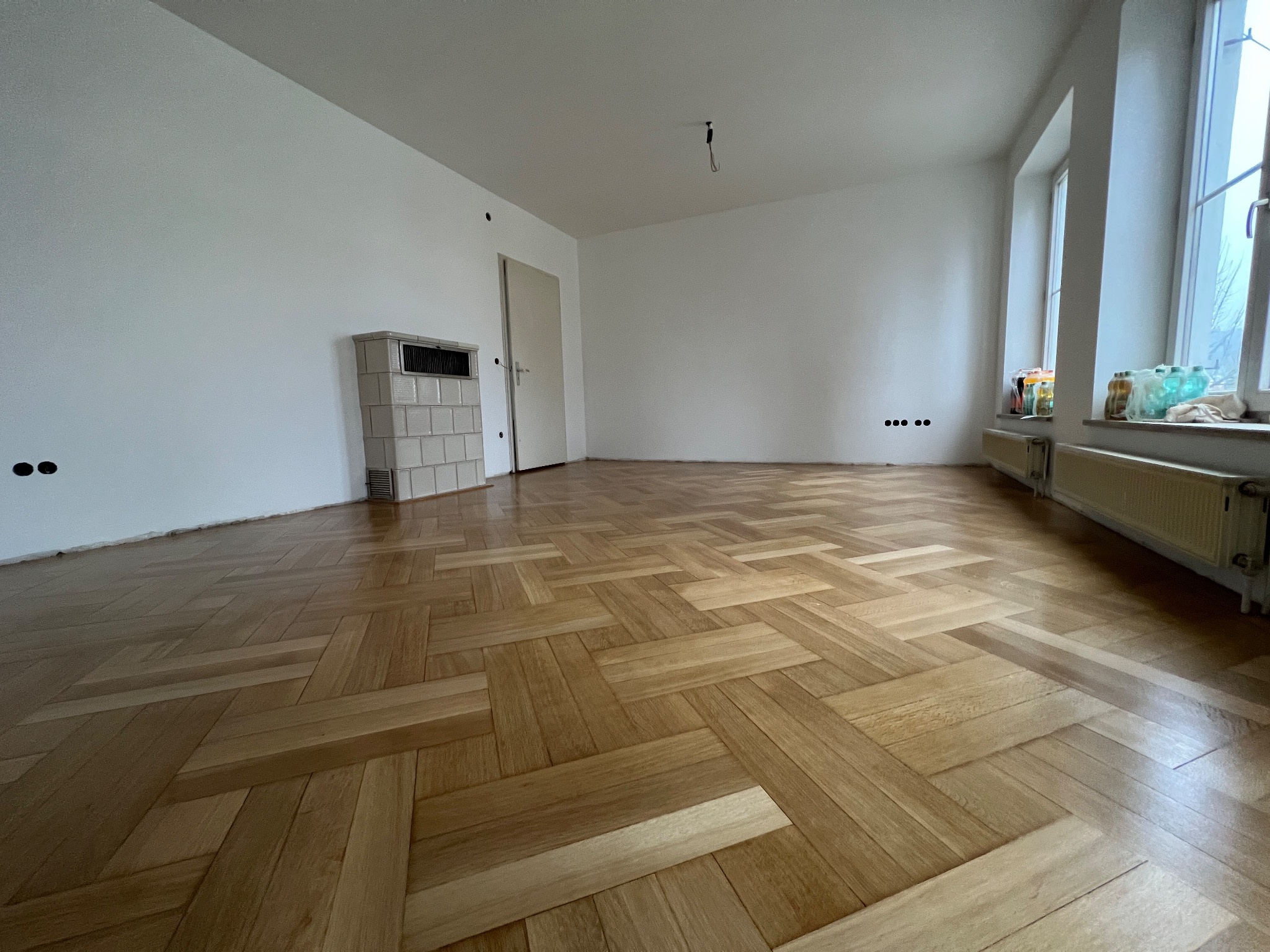 Jasne, nowo ułożone panele podłogowe w jodełkę w pustym, odnowionym pokoju z białymi ścianami i grzejnikiem pod oknem.