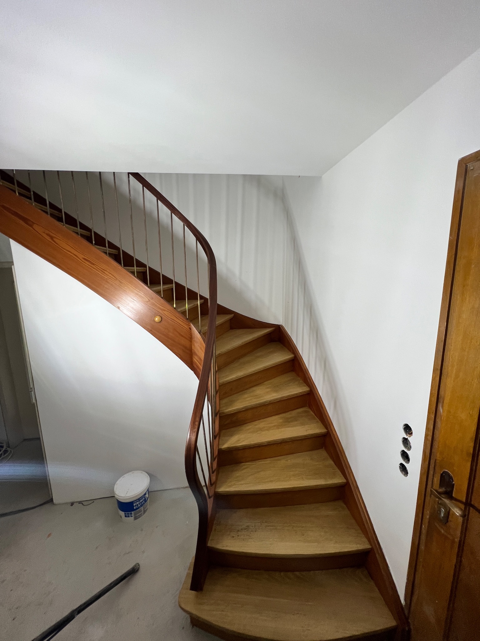 Drewniane, kręte schody z metalową balustradą, prowadzące na górę w nowo pomalowanym, jasnym pomieszczeniu. Widoczna puszka z farbą na podłodze i niezamaskowane gniazdka elektryczne.