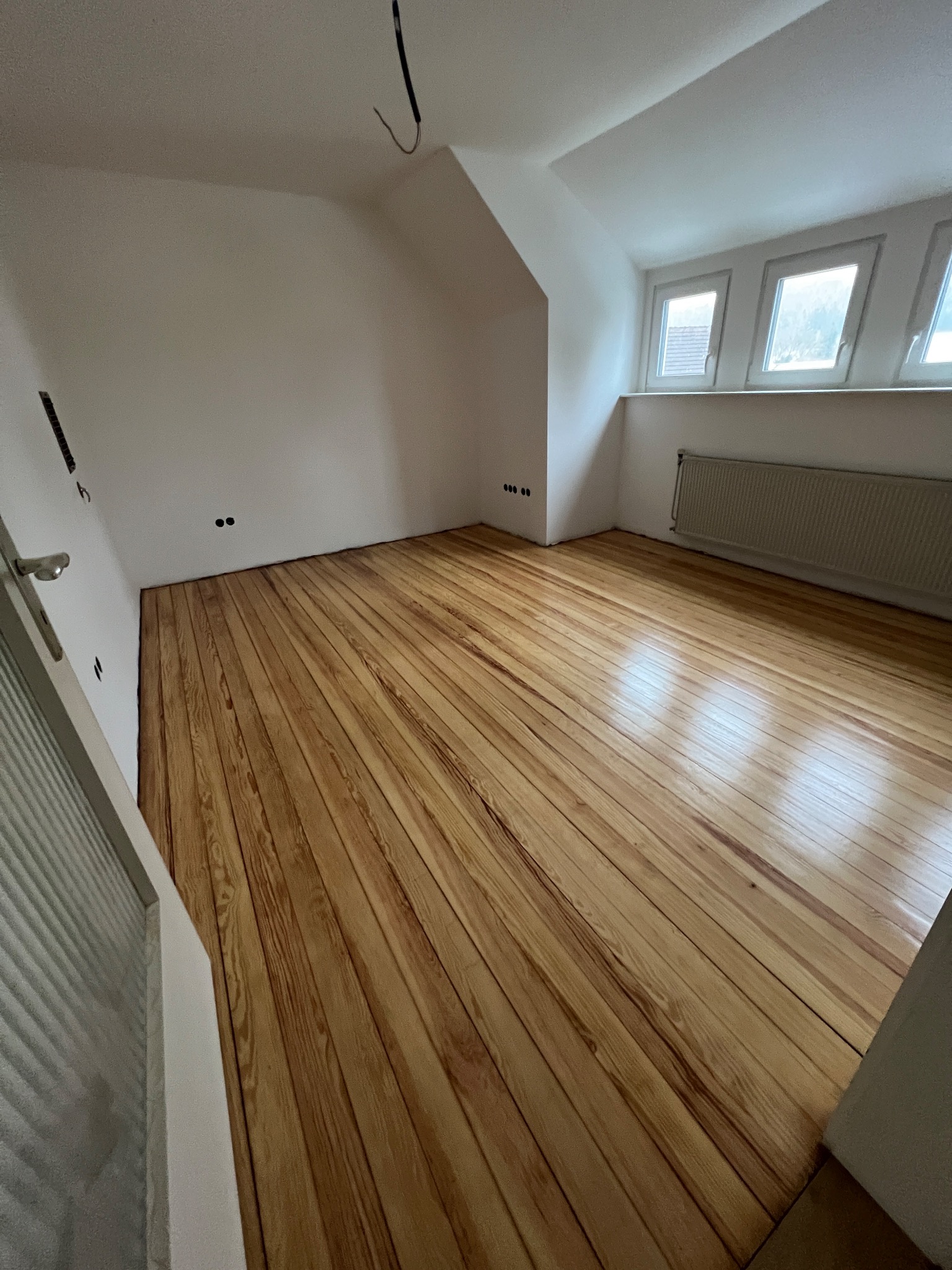 Poddasze z nowo ułożoną, lakierowaną podłogą z desek o ciepłym odcieniu, widoczne okna dachowe i grzejnik.
