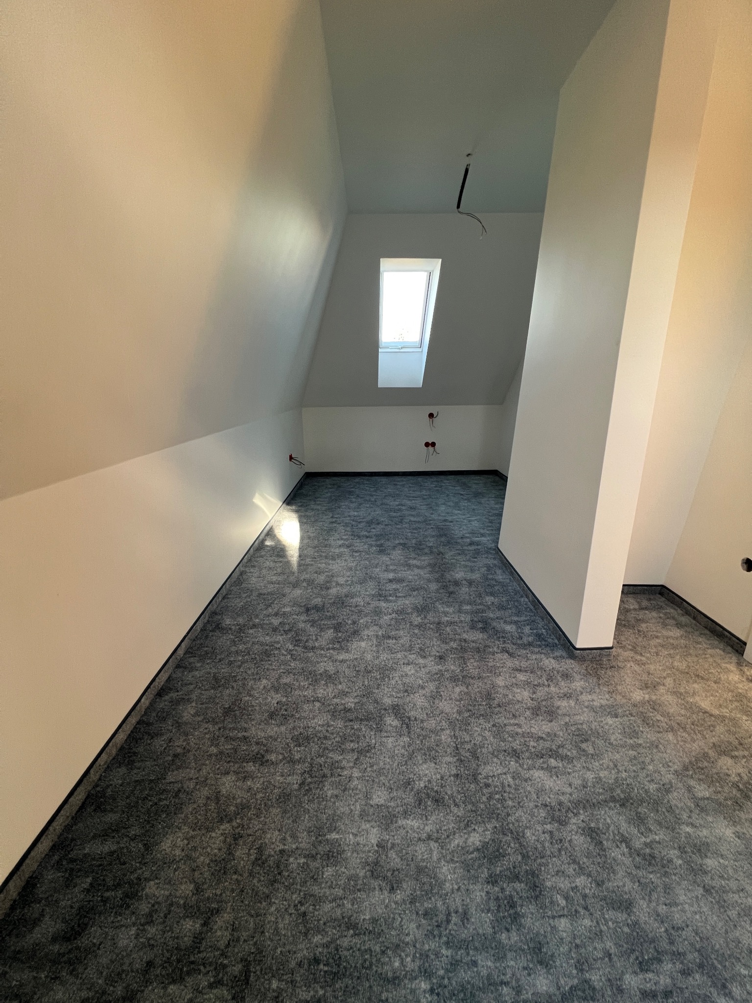 Poddasze po remoncie z nową szarą wykładziną dywanową, widoczne gładkie, pomalowane na biało ściany ze skosami i oknem dachowym.