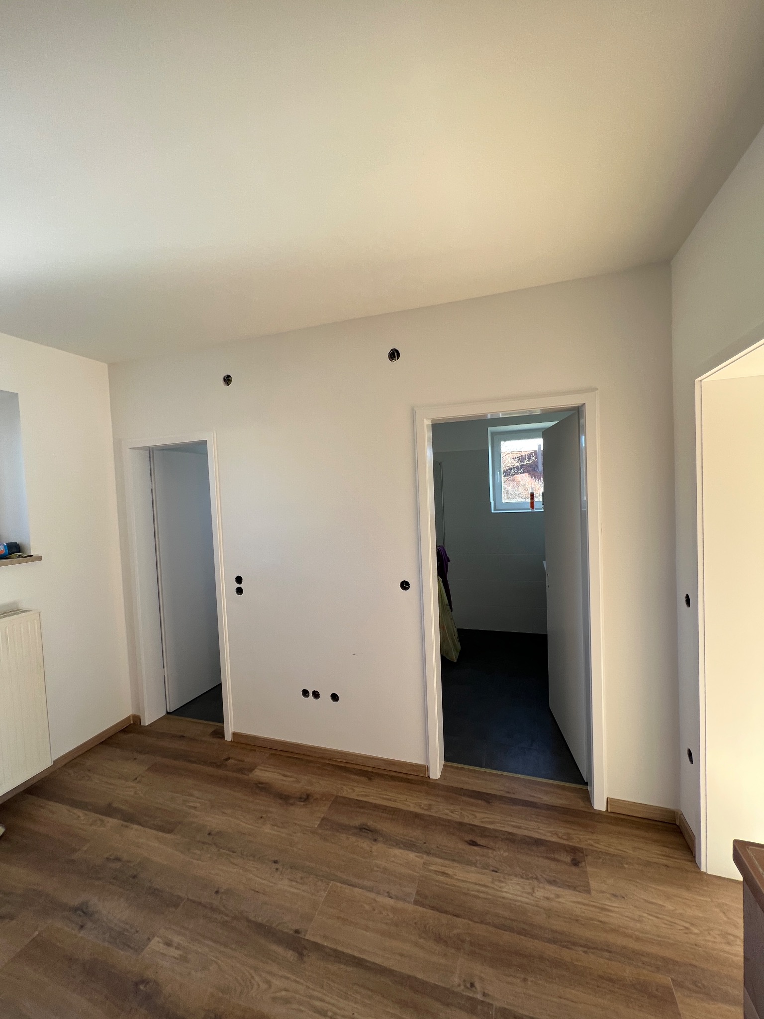 Jasne wnętrze pokoju po remoncie, z ułożonymi panelami podłogowymi w odcieniach brązu, świeżo pomalowanymi na biało ścianami z widocznymi otworami po instalacjach i dwoma otwartymi przejściami...