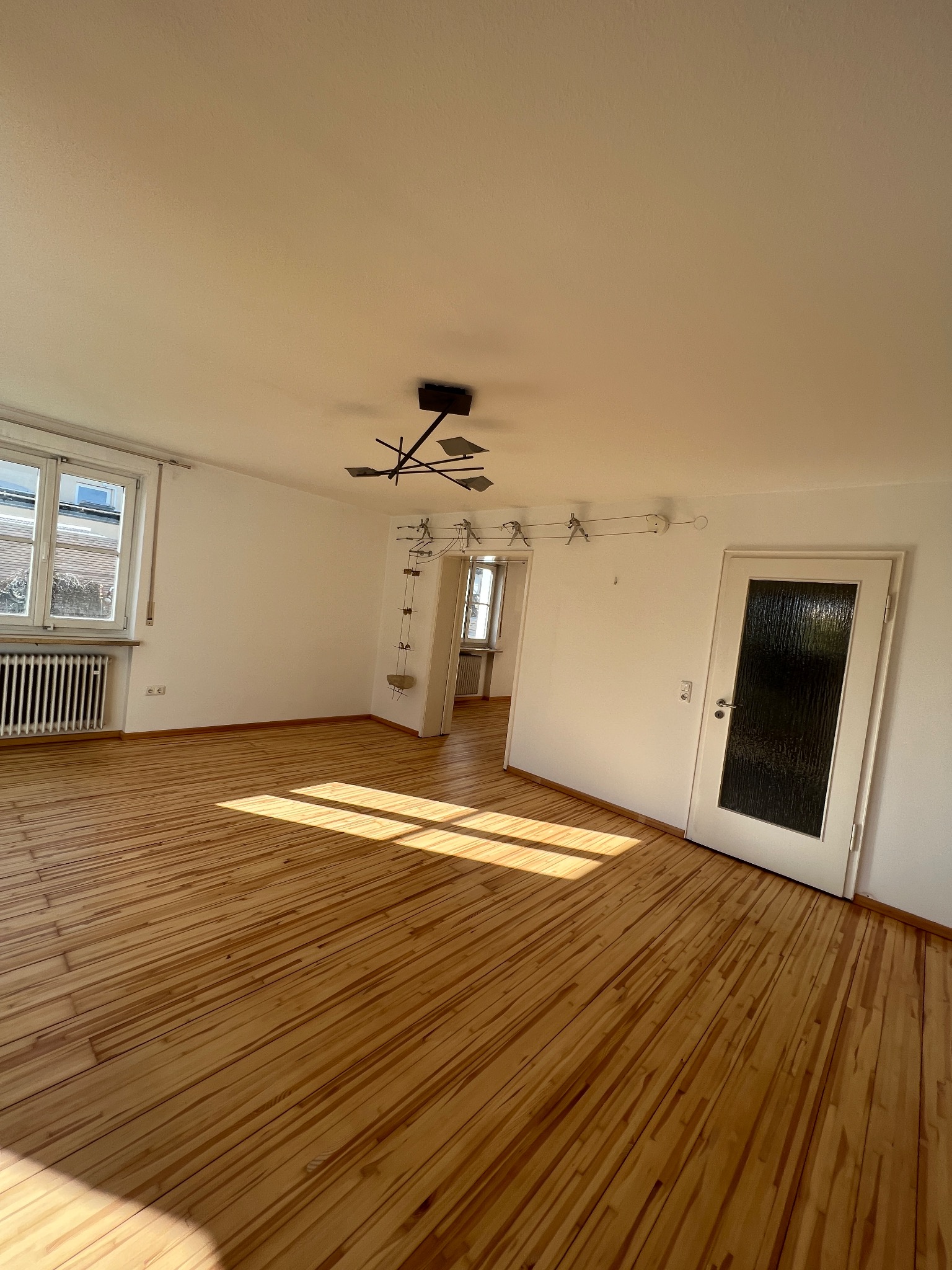 Jasne, puste pomieszczenie z drewnianą podłogą panelową, oświetlone promieniami słonecznymi wpadającymi przez okno. Widoczny minimalistyczny żyrandol i białe ściany.