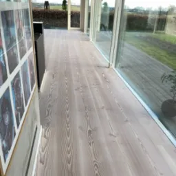 Jasna, odnowiona drewniana podłoga z wyraźnym usłojeniem w nowoczesnym wnętrzu z dużymi oknami i widokiem na zewnątrz, z dekoracyjnym obrazem na ścianie po lewej stronie.