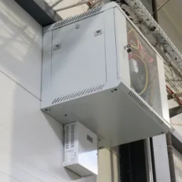 Szara szafa sterownicza z przeszklonymi drzwiczkami, zamontowana na ścianie hali, widoczne przewody wewnątrz, dodatkowy moduł z radiatorem poniżej, koryta kablowe nad szafą.