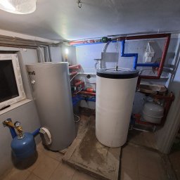 ARKOS - INSTAL ARKADIUSZ STENKA - Dwa zbiorniki na wodę, jeden szary marki Ferroli, drugi biały, połączone rurami w pomieszczeniu kotłowni z widocznymi elementami instalacji grzewczej.